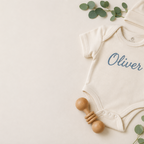 Personalized cotton baby bodysuit embroidered with the name Oliver, flat lay with knit hat and wooden rattle